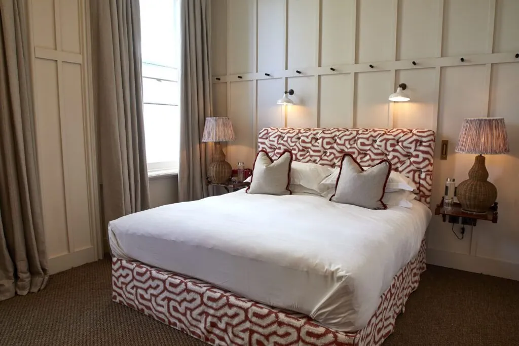 A bedroom above The Princess Royal with white panelled walls, a buttoned headboard and bed base in red and white geometric fabric, wicker lamps on wall-hung shelves and a tall window with linen curtains
