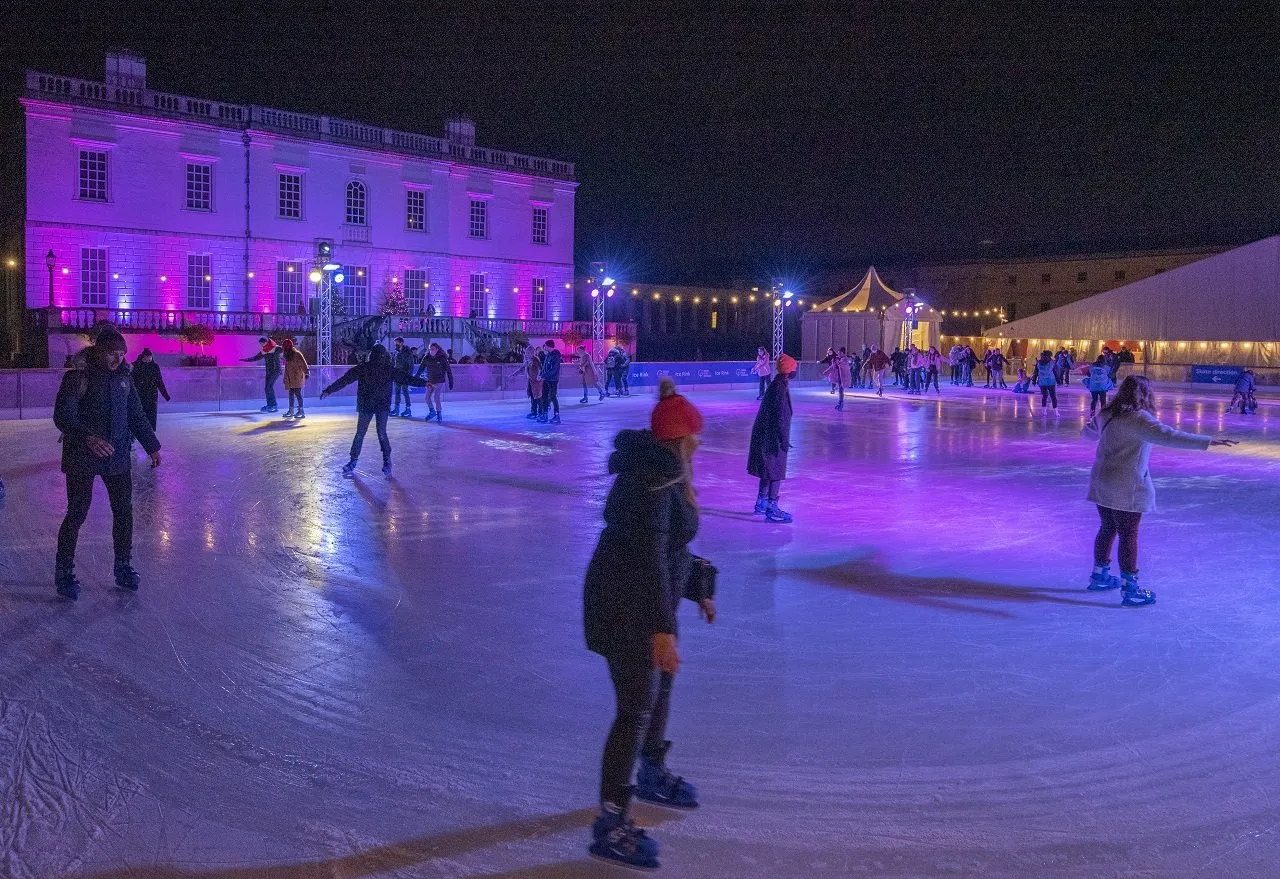 Skaters on the Queen's House ice rink in Greenwich at night, the white Palladian villa lit pink and purple behind them and a marquee to the right