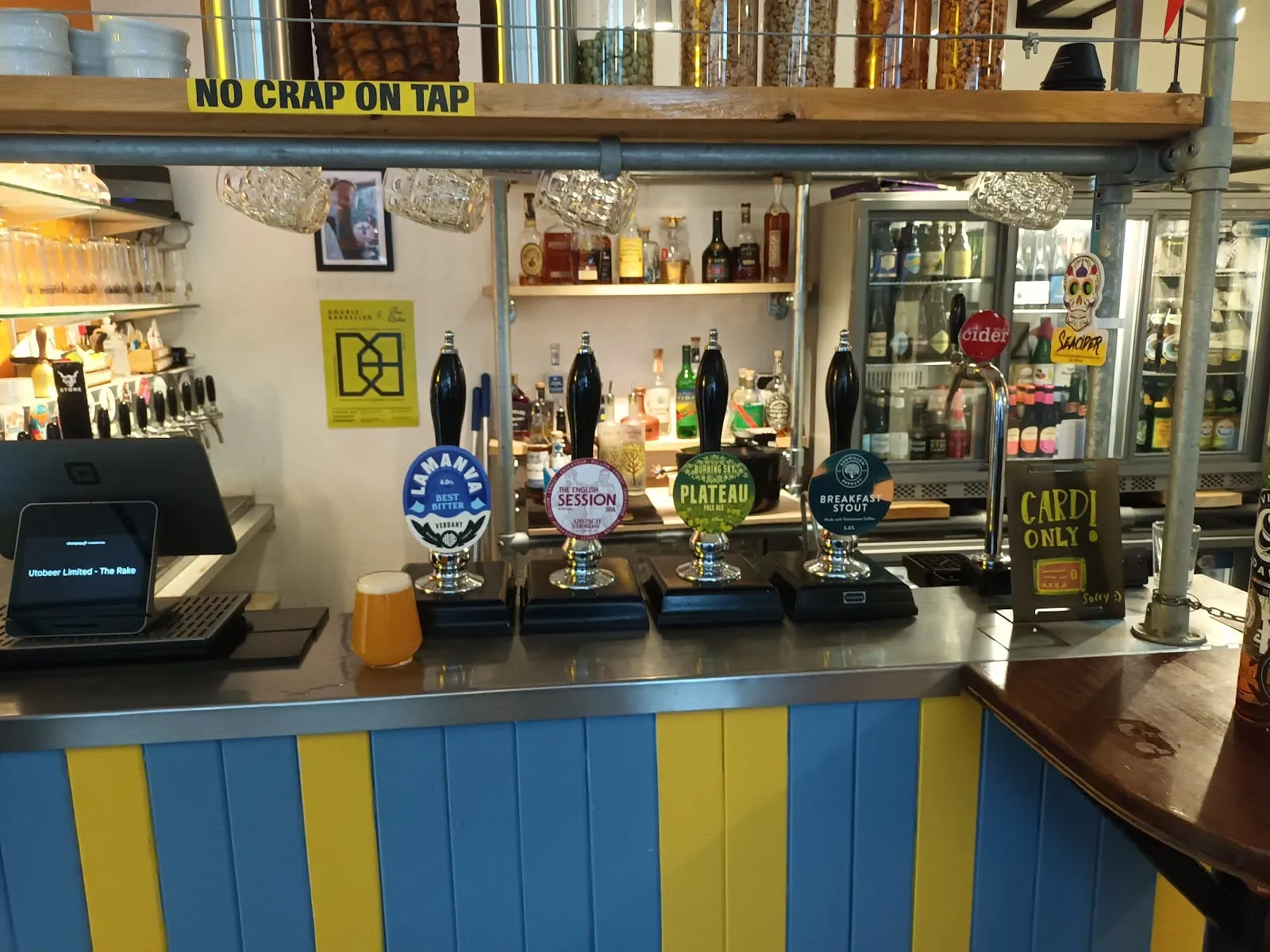 Handpumps at The Rake below a sign reading "No crap on tap", with fridges of bottles behind the bar