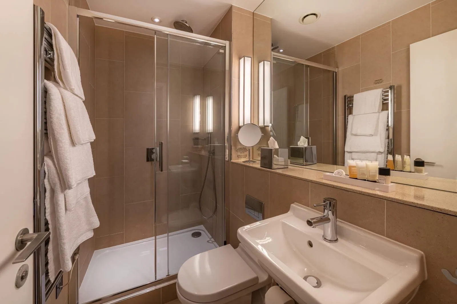 A bathroom at The Resident Victoria in beige stone tiles, with a shower behind sliding glass doors, a heated towel rail and a white basin