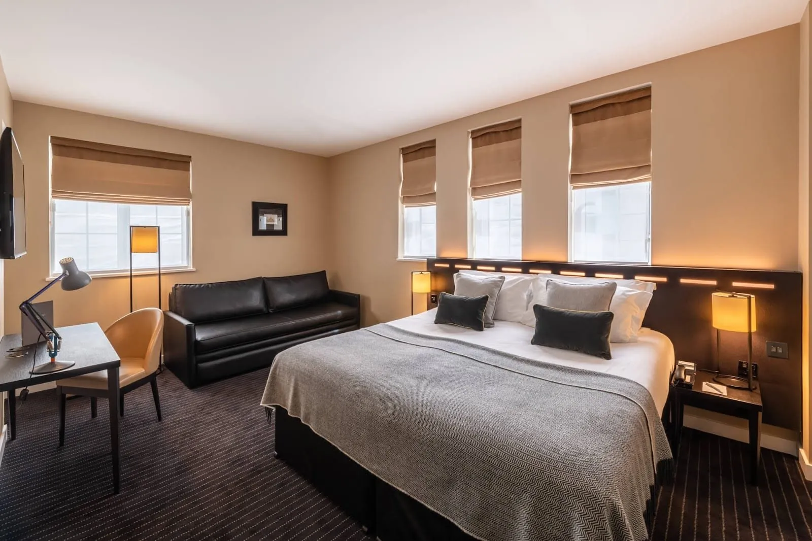 A room at The Resident Victoria with a grey herringbone throw, a backlit headboard under three windows with roman blinds, a black leather sofa and a desk with an anglepoise lamp