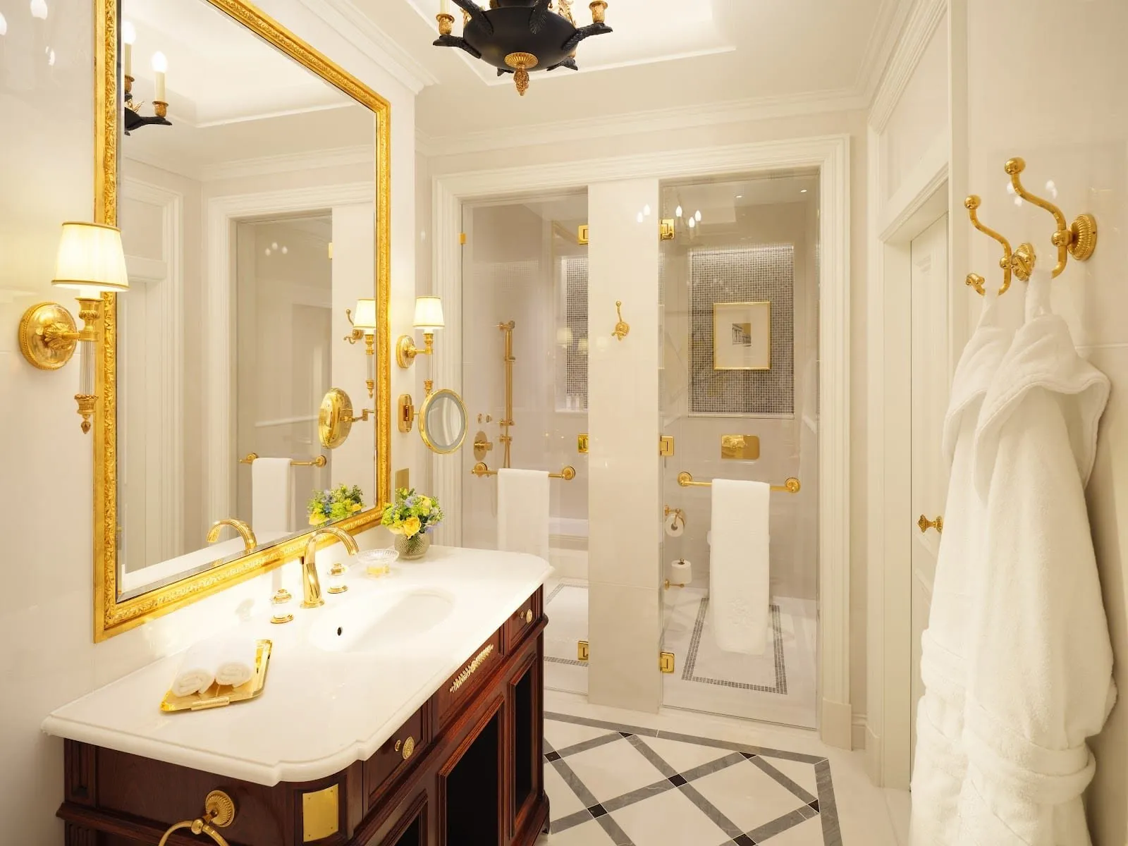 A Ritz bathroom in cream marble with a gilt-framed mirror, gold taps on a mahogany vanity, a glass-walled shower, a separate WC and two white bathrobes on brass hooks