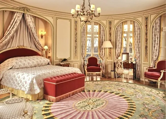 An opulent Louis XVI-style suite at The Ritz London with a draped bed canopy, crystal chandelier and circular patterned rug