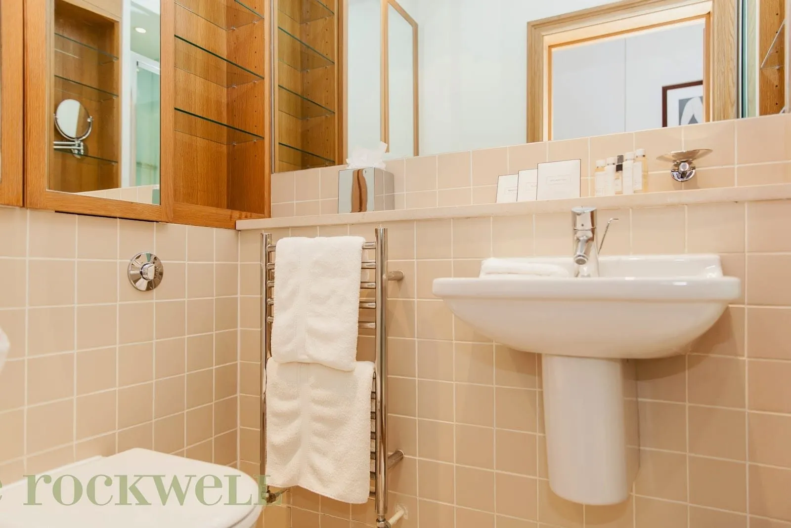 A Rockwell bathroom in peach tiles with a wall-hung basin, a heated towel rail and oak-framed mirrored cabinets with glass shelves