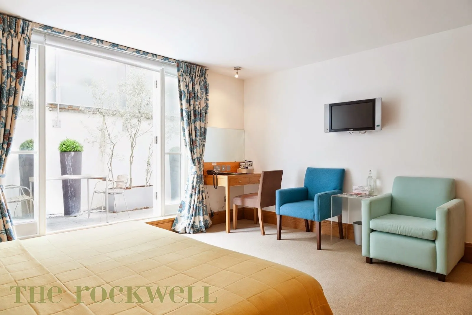 A Garden Room at The Rockwell with a mustard bedspread, teal and blue armchairs and a desk, and full-height glass doors opening onto a private patio with a table, chairs and olive trees