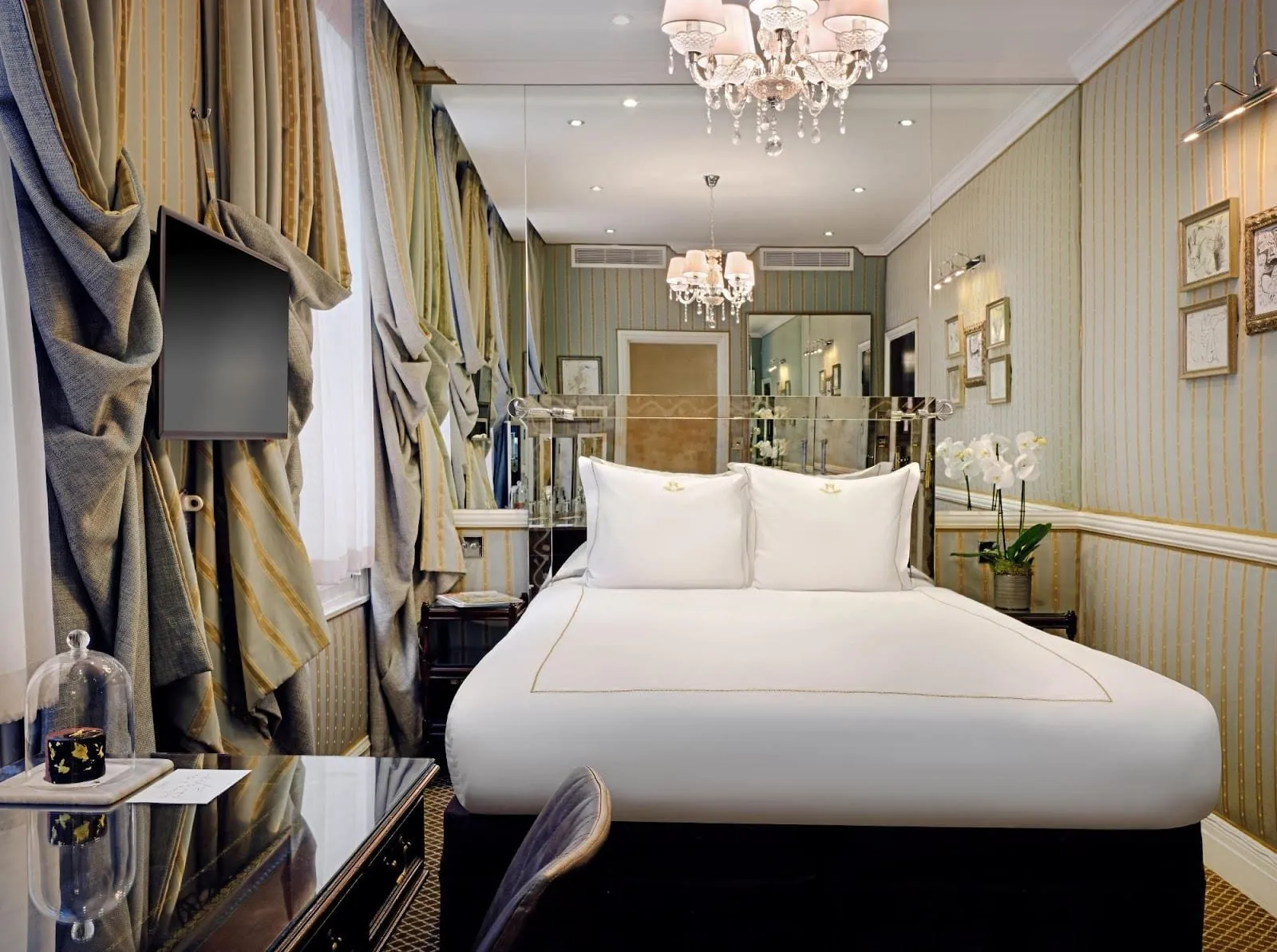 A narrow double room at The Rubens at the Palace, with a crystal chandelier, a mirrored wall behind the bed, striped silk wallpaper and heavy swagged curtains beside a wall-mounted TV