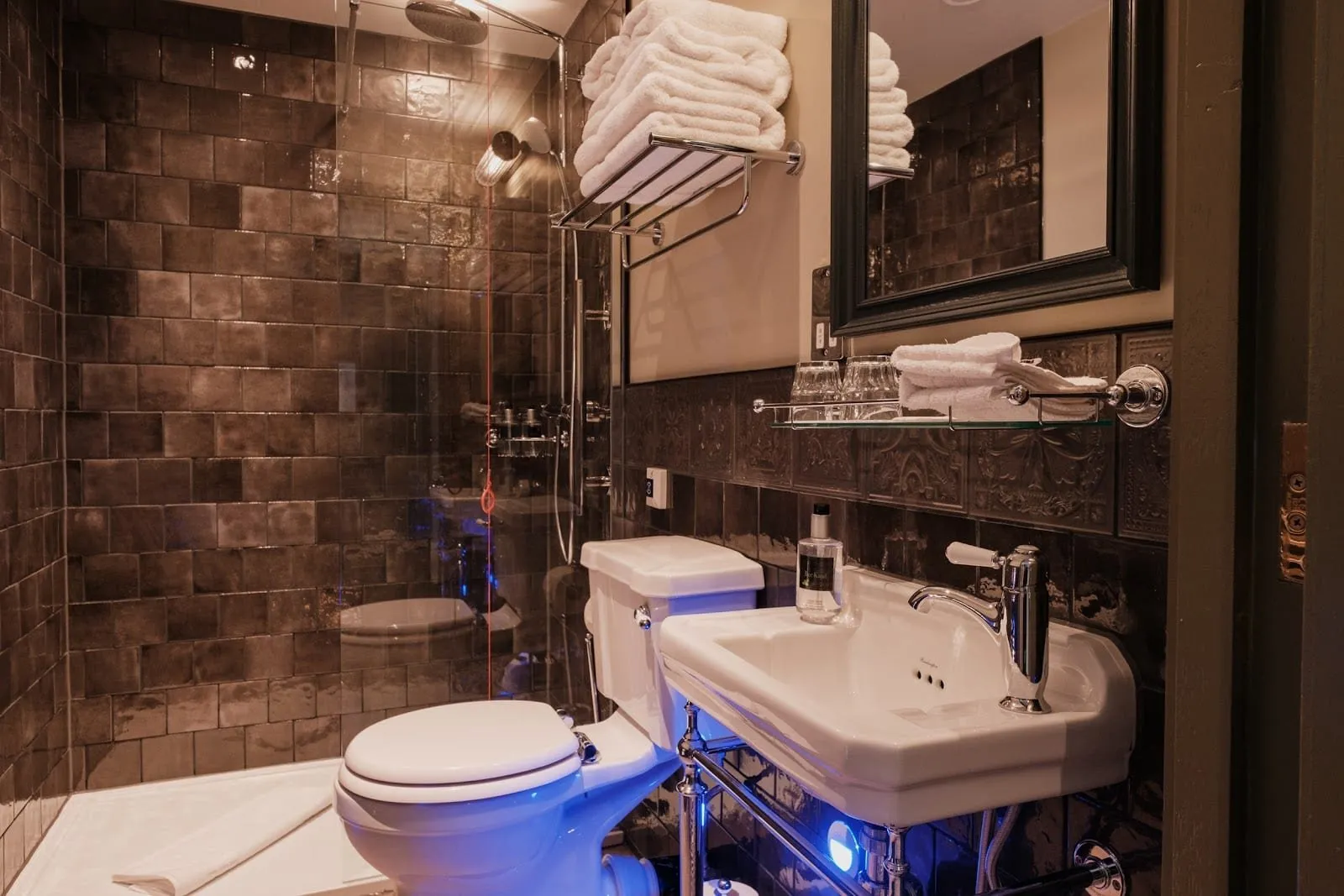 A bathroom at The Sanctuary House Hotel with dark glazed tiles, a walk-in rain shower behind glass, a Victorian-style basin on a chrome stand and a towel rack above the toilet