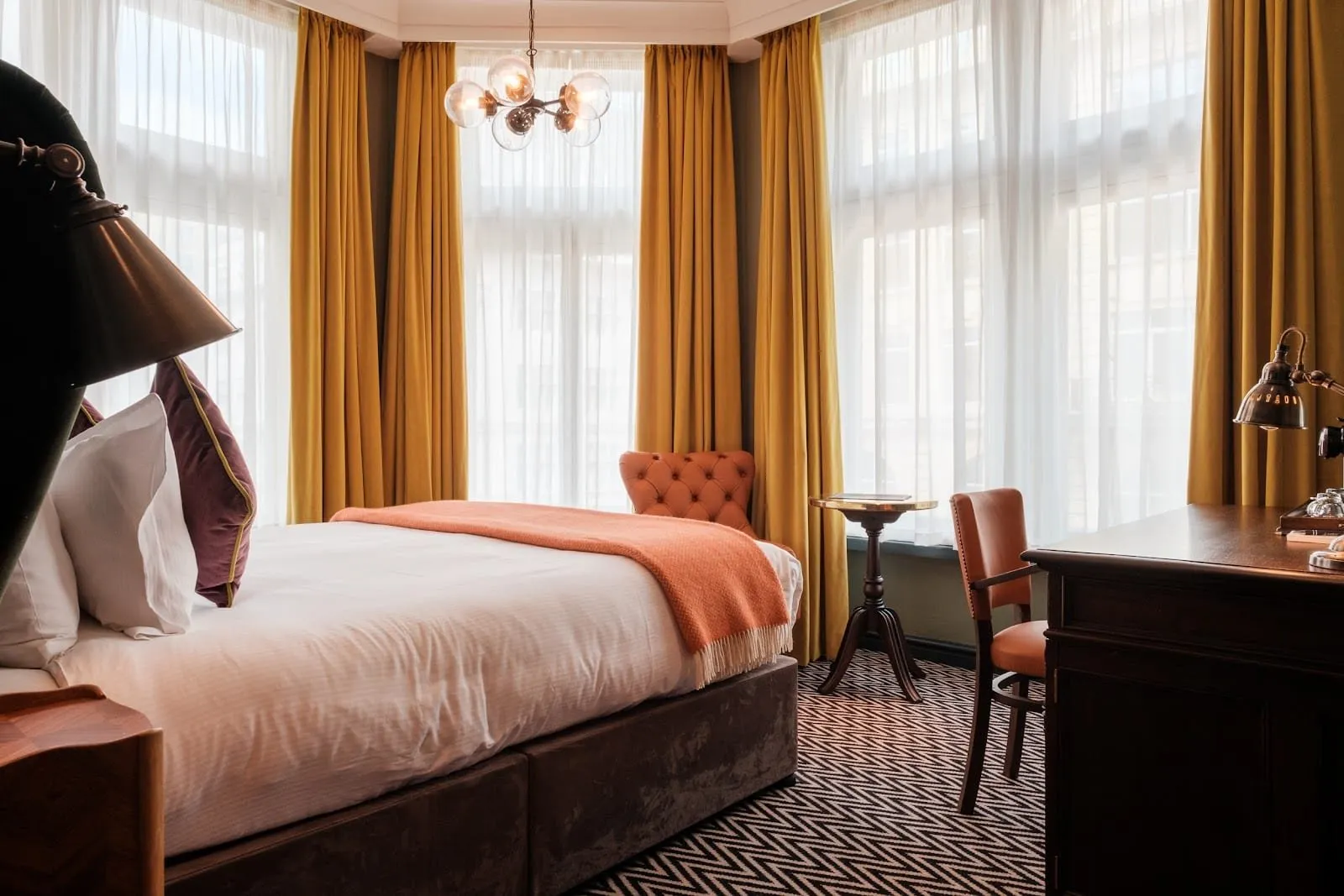 A room at The Sanctuary House Hotel with a bay window hung with mustard velvet curtains, a bed with an orange throw, a glass globe chandelier, a chevron carpet and a wooden desk with a brass lamp
