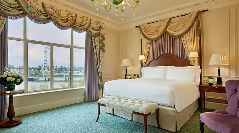 A guest room at the Savoy, with heavy drapery, a carved wood headboard and the Thames and London Eye visible through the window