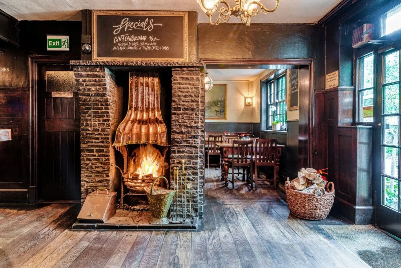 A lit fire under a copper hood in a brick fireplace at The Spaniards Inn, with a dining room visible through the doorway beyond