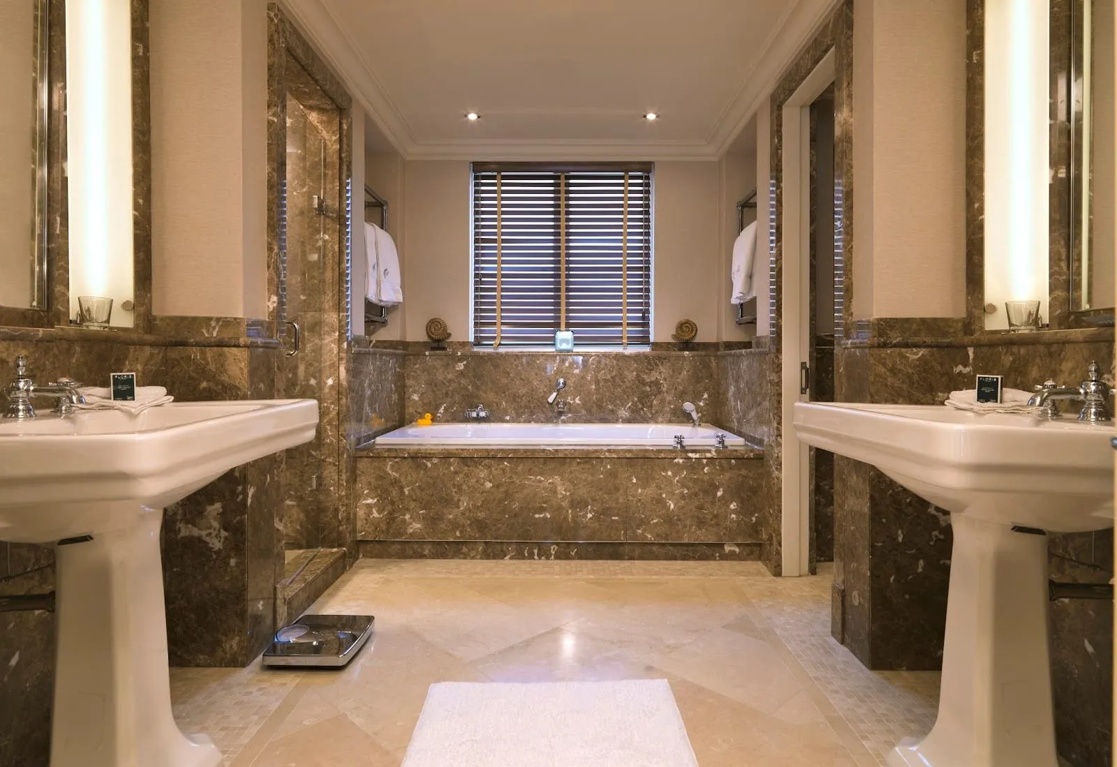A bathroom at The Stafford in brown marble, with two pedestal basins facing each other, a bath under a window with wooden blinds and a separate glass shower