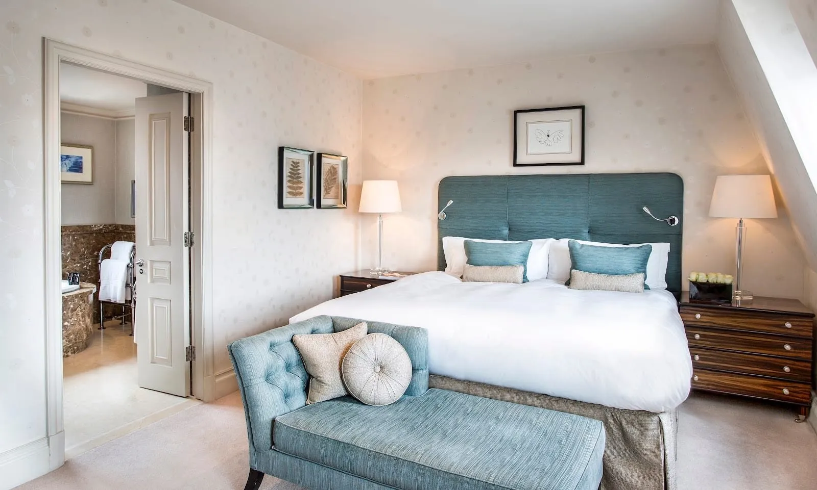 A room at The Stafford under a sloping ceiling, with a teal padded headboard, a teal chaise at the foot of the bed and a door open to a marble bathroom