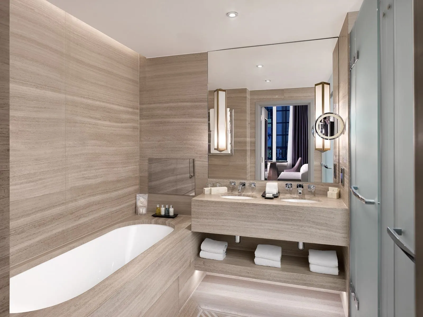 A bathroom at The Trafalgar St. James in pale striped stone, with a bath, twin basins under a wide mirror and a frosted glass shower door