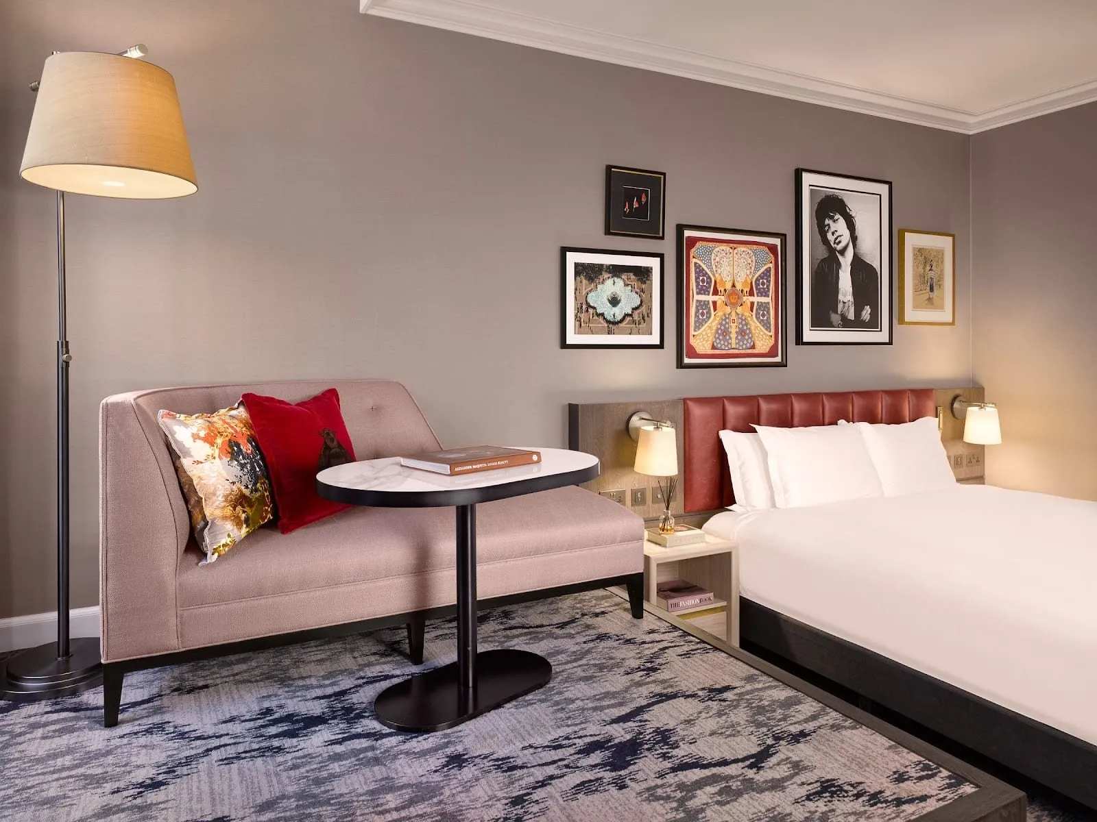 A room at The Trafalgar St. James with a red leather headboard, a pink chaise and small marble table, and a gallery wall of prints including a black-and-white portrait photograph