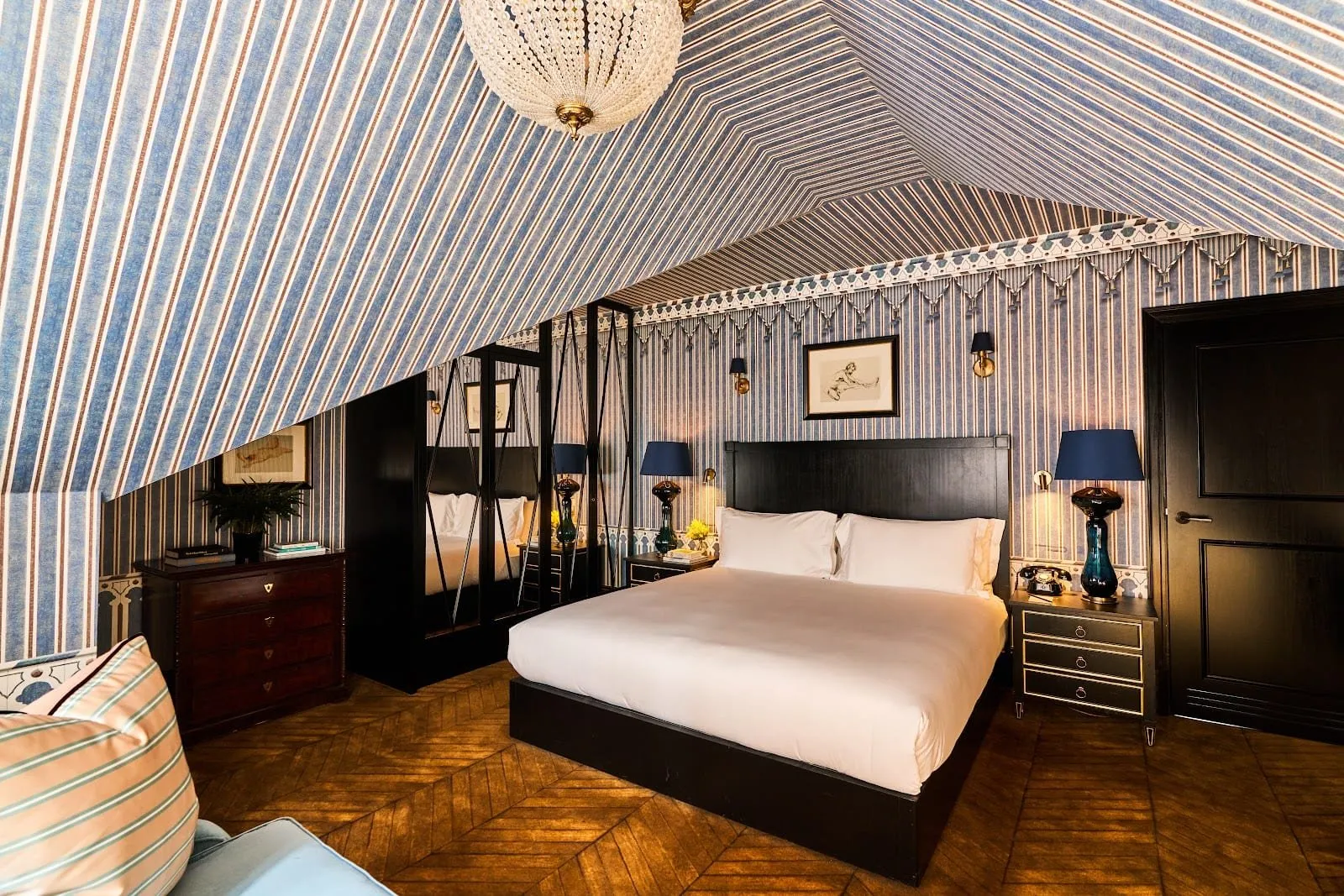 A top-floor Twenty Two bedroom under a sloping ceiling covered in blue and white striped fabric like a tent, with a beaded chandelier, a black-framed double bed, navy lampshades and a herringbone wood floor