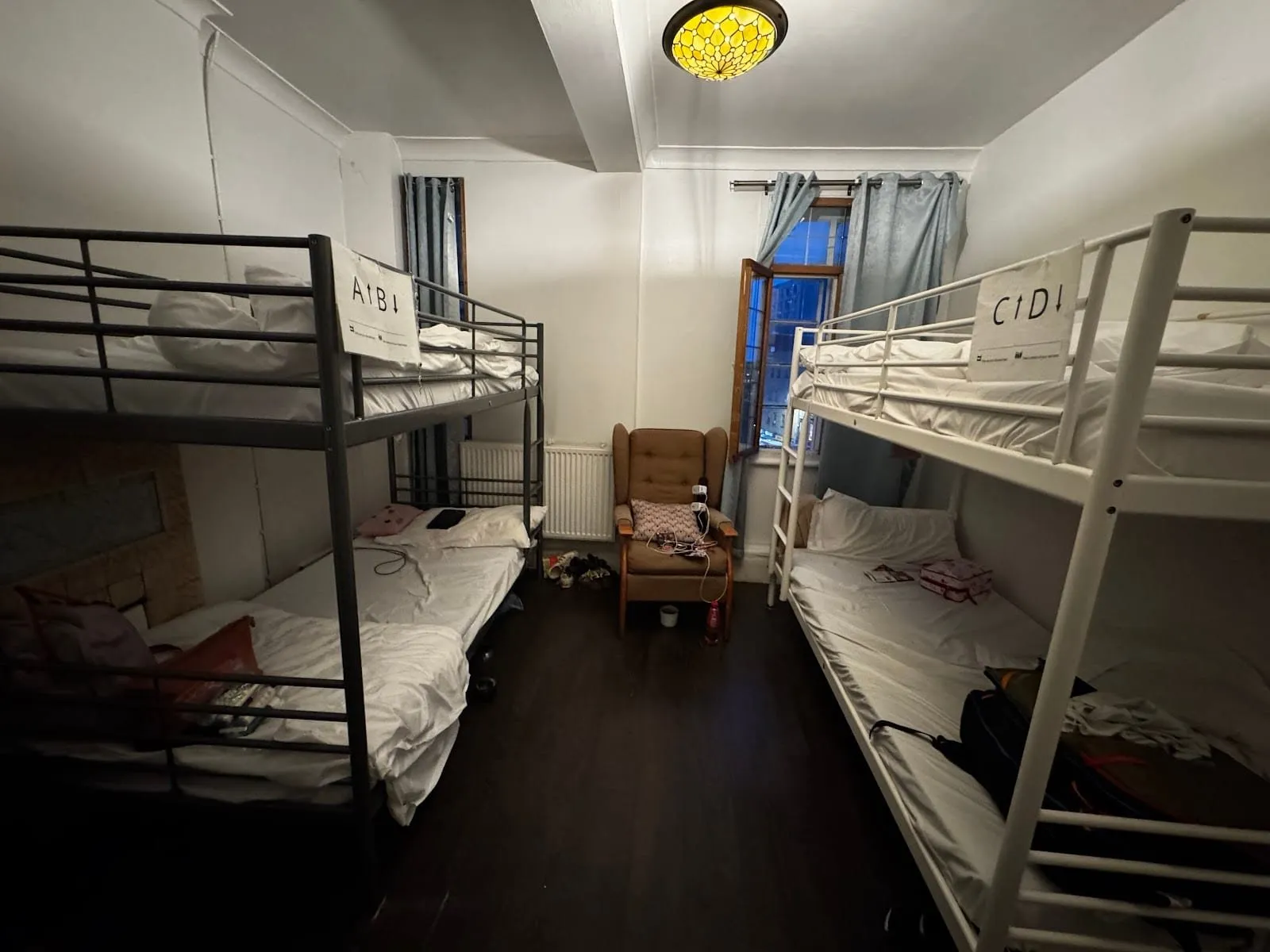 A four-bed dorm at The Walrus with two metal bunk beds labelled A to D, a dark wooden floor, a brown armchair and an open window at night