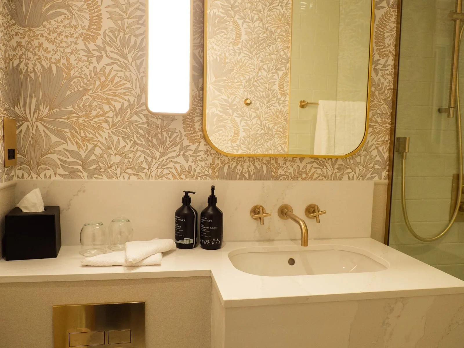 A bathroom at The Welbeck with gold and grey leaf wallpaper, a white quartz basin with brushed brass taps, a round-cornered brass mirror and a glass-screened shower on the right