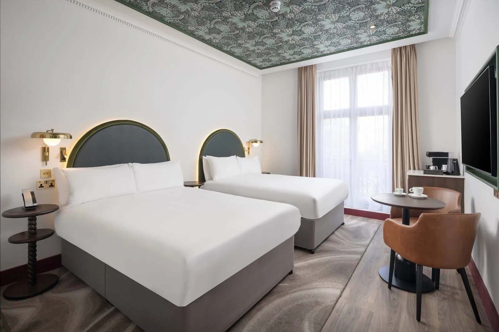 A room at The Welbeck with two double beds under arched green headboards, brass wall lamps, a leaf-print ceiling panel, two leather tub chairs at a small table and tall French windows with an iron balcony rail