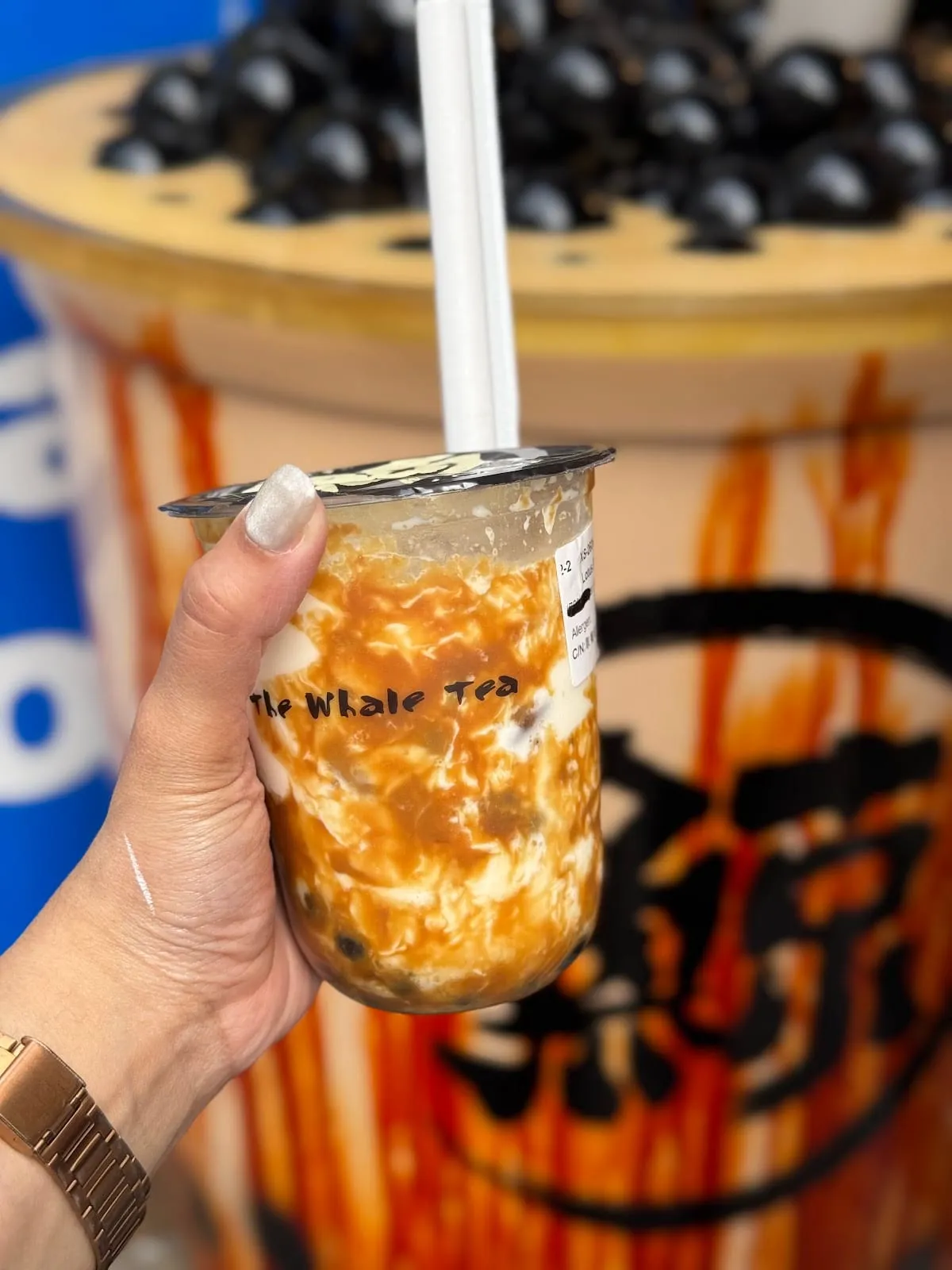 A clear cup of brown sugar boba streaked with caramel syrup and cream, held up in front of a larger cup piled with pearls