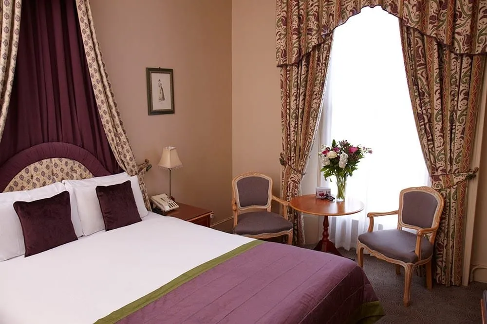 A double room at The Windermere with a plum and gold fabric canopy over the bed, purple velvet cushions, and two carved chairs at a round table in a tall window with patterned swagged curtains