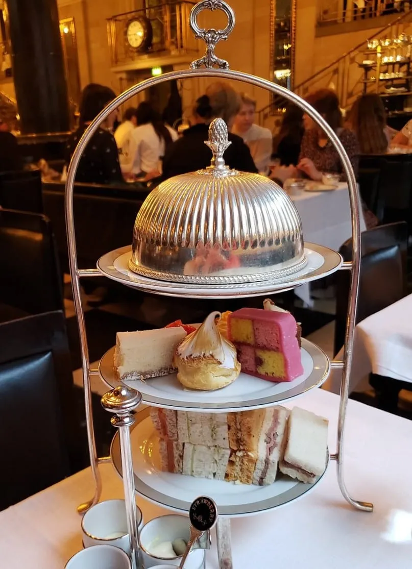 A tiered stand under a silver cloche holding battenberg, cheesecake and finger sandwiches, in the Wolseley's vaulted room with the clock behind
