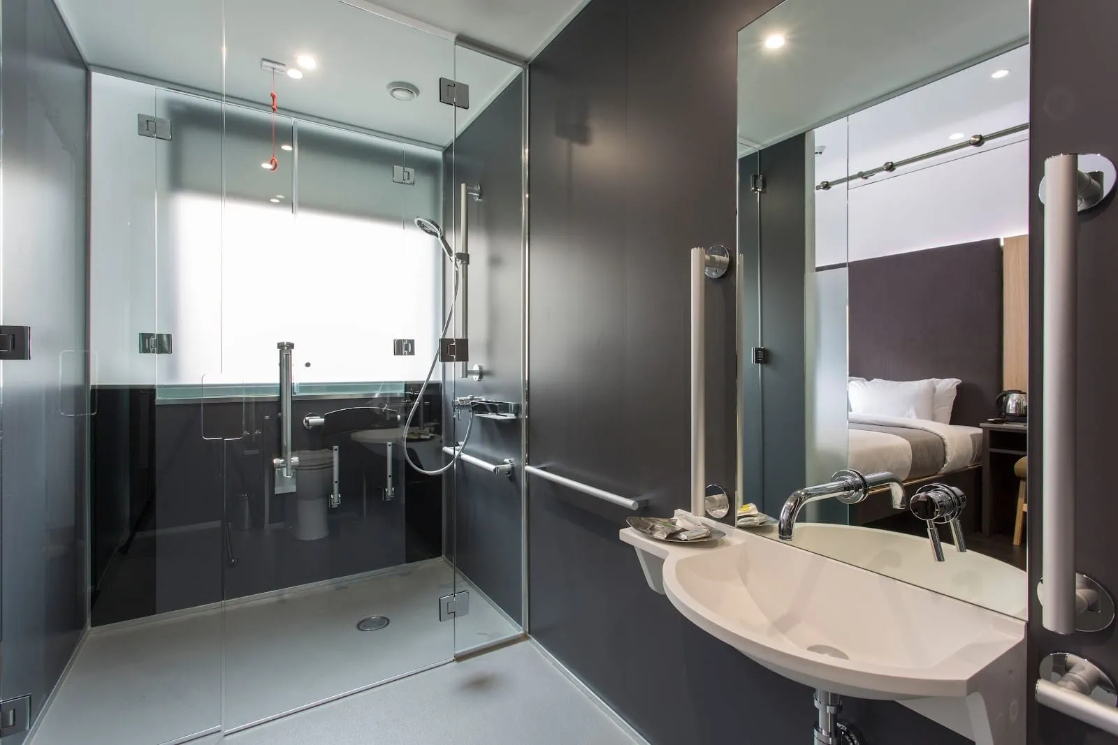 The accessible wet room at The Z Hotel City: a glass-walled walk-in shower with a red alarm cord, grab rails around the toilet and a low white basin under a wide mirror