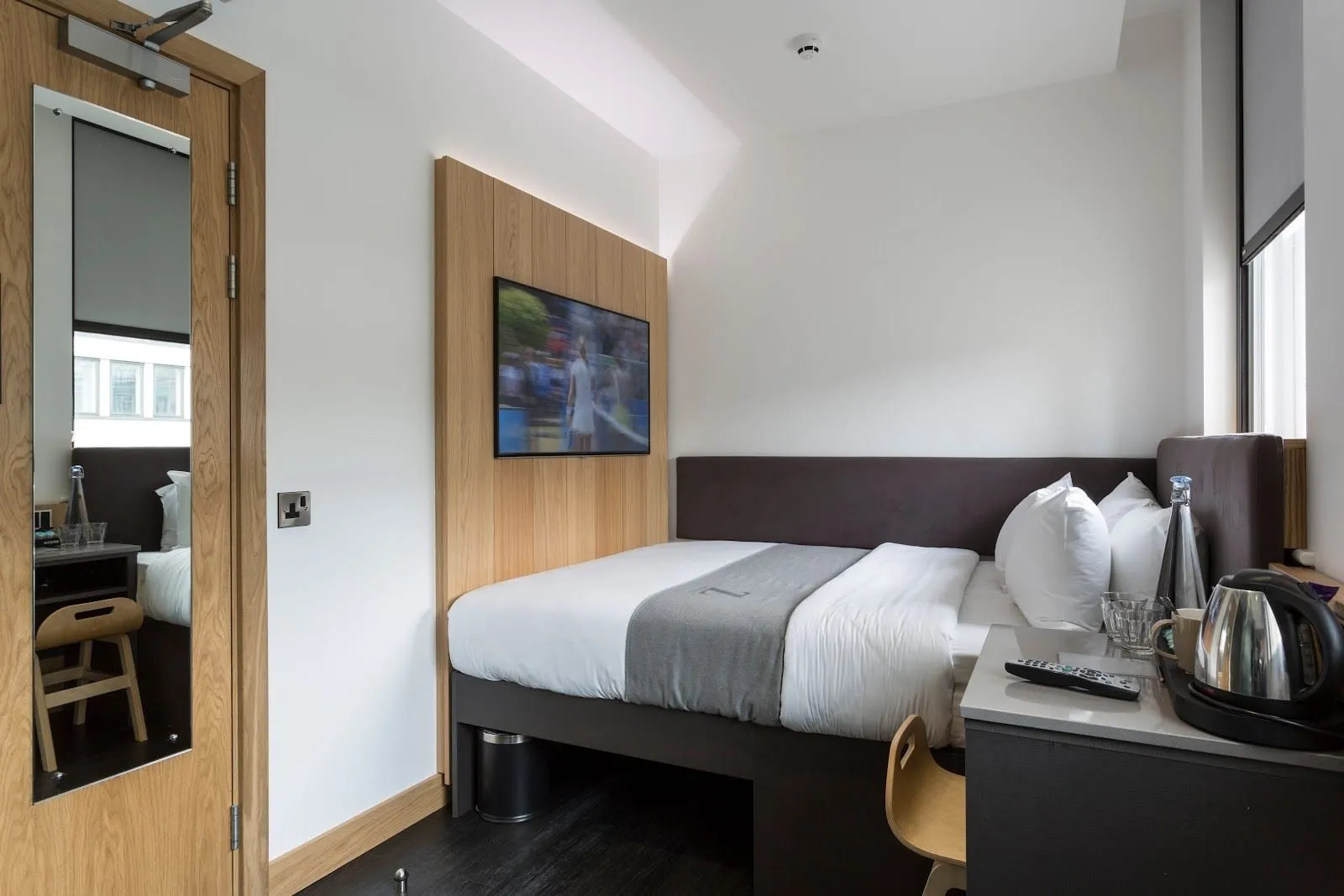 A compact double at The Z Hotel City filling the width of the room against a charcoal padded headboard, with a TV on an oak panel, a small desk with a kettle, and a window with a blind beside the bed