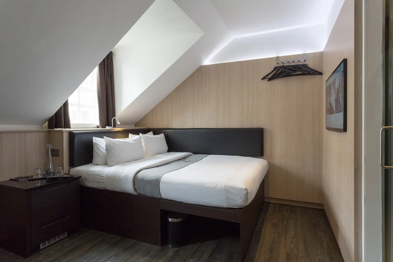 A room at The Z Hotel Victoria under a sloping attic ceiling, with a double bed filling the space between wood-panelled walls, a black padded headboard, a dormer window beside the head of the bed and a wall-mounted TV