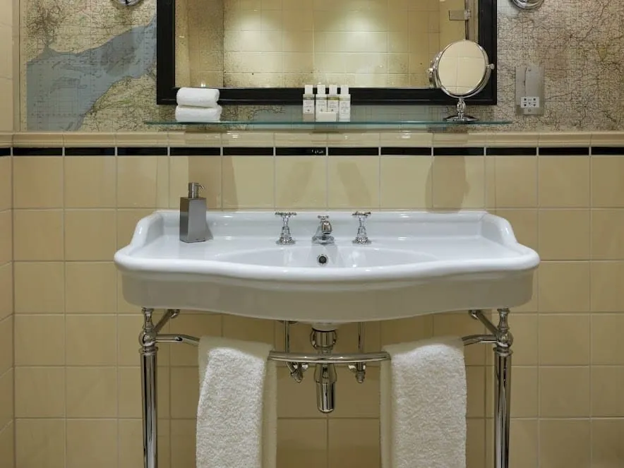 A bathroom at The Zetter Marylebone with a white washstand basin on chrome legs, cream wall tiles with a black border and old map wallpaper around the mirror