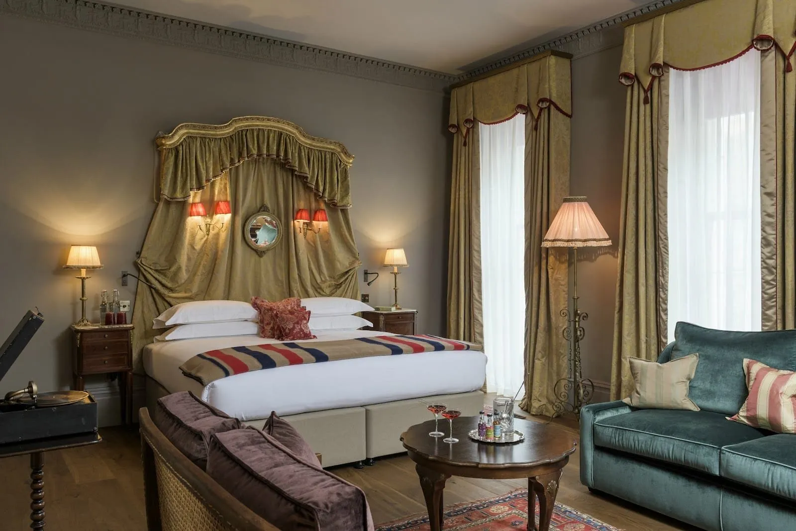 A room at The Zetter Marylebone with olive drapes hung in a gilded canopy behind the bed, red-shaded wall lights, a teal velvet sofa, a wind-up gramophone and two tall windows with swagged pelmets