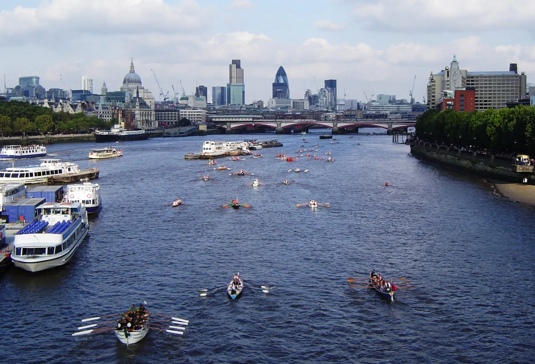 Dozens of traditional rowing boats spread across the River Thames during the Great River Race, with the dome of St Paul's Cathedral and the City skyline behind, Blackfriars Bridge in the middle distance and spectators lining the embankment