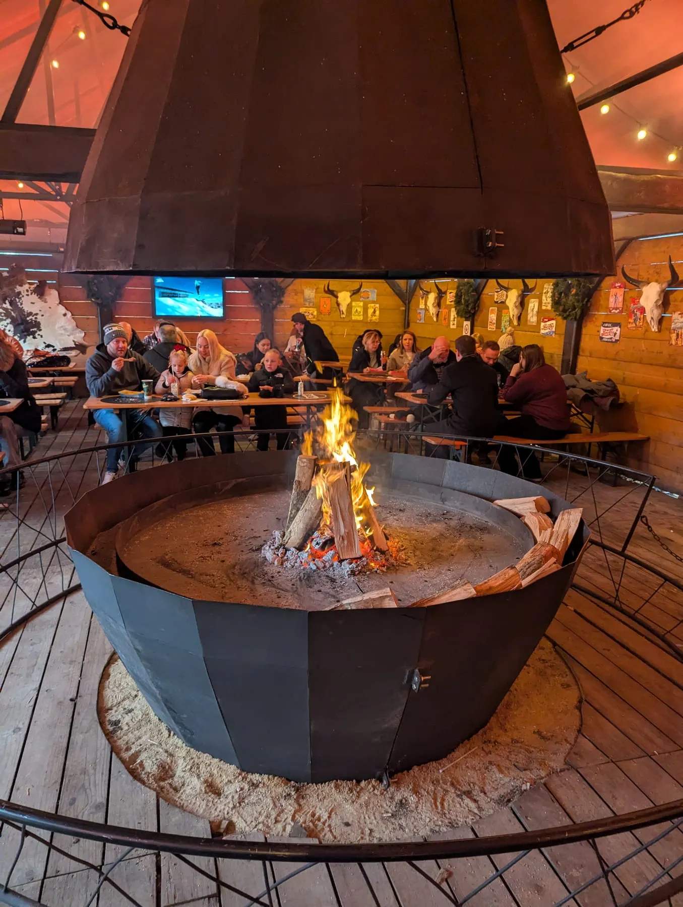 Inside Thor's Tipi Bar at Winter Wonderland: a large open fire burning in a circular steel pit under a metal hood, with people eating at long wooden benches around the edge