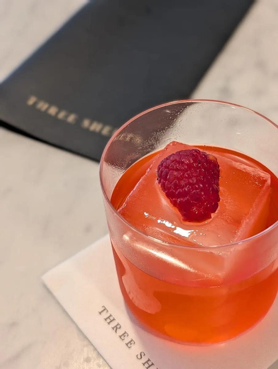A pink drink over one large ice cube with a raspberry on top, on a Three Sheets napkin next to a black menu on a marble counter