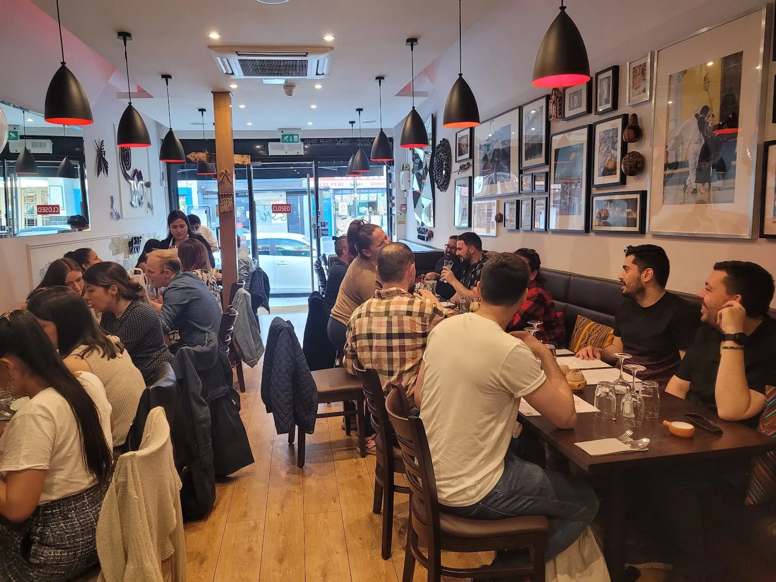 A small, full restaurant with white walls, black pendant lamps, Peruvian textile cushions on the banquette and a dense gallery wall of framed photographs, every table occupied