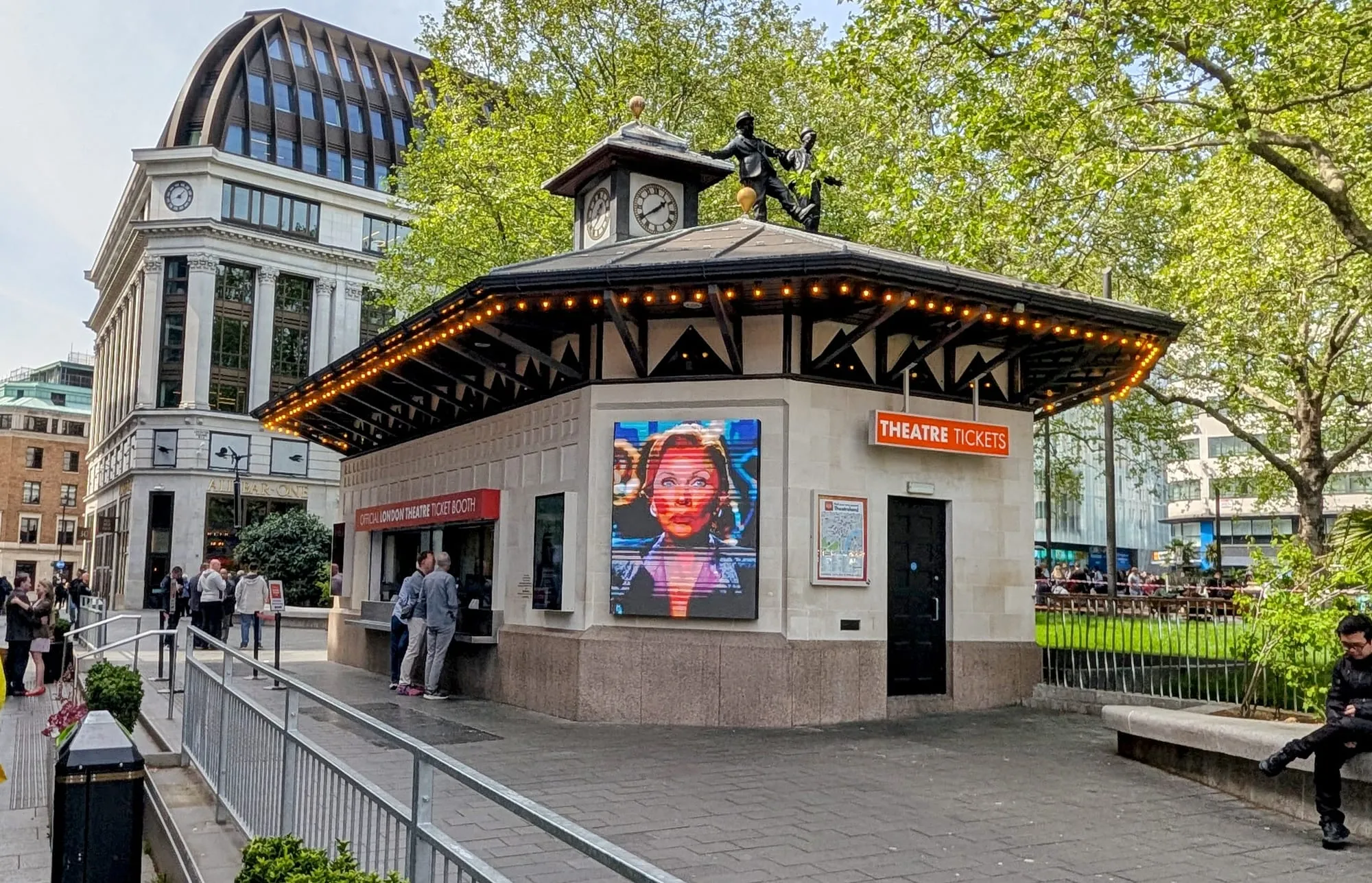 The clocktower building of the official TKTS theatre ticket booth in Leicester Square, with people queuing at the "OFFICIAL LONDON THEATRE TICKET BOOTH" window