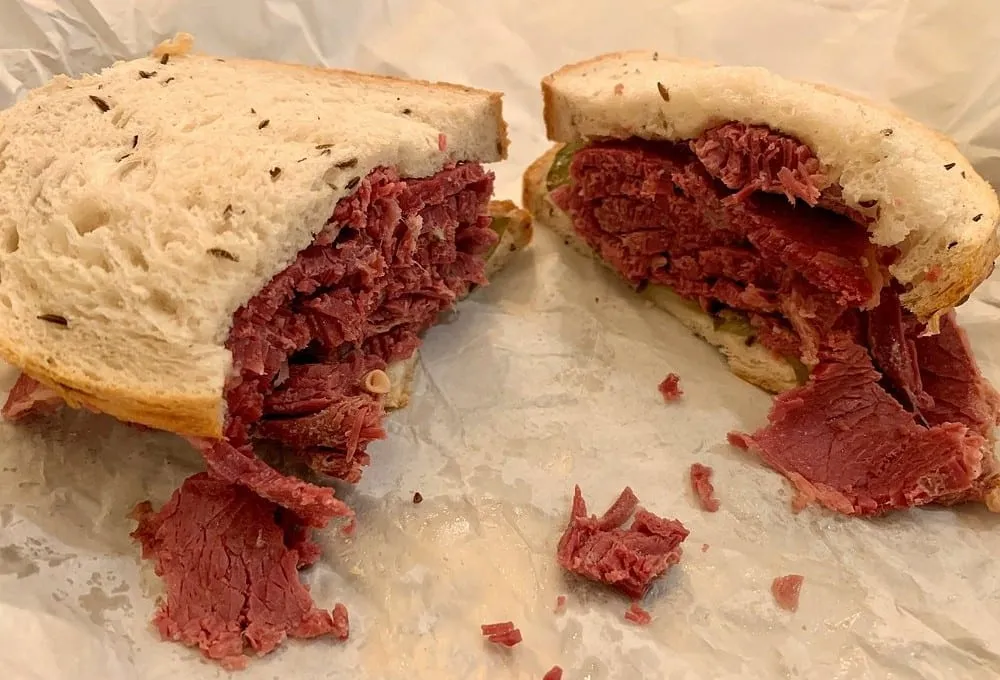 A salt beef sandwich cut in half on greaseproof paper, thick hand-carved beef spilling out of caraway-seeded bread with pickle showing at the back