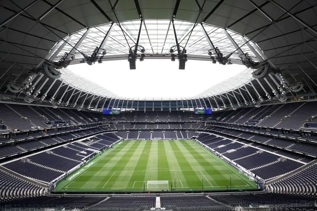 Tottenham Hotspur Stadium's bowl seen from a high tier, the retractable roof open above rows of navy seating and an empty pitch