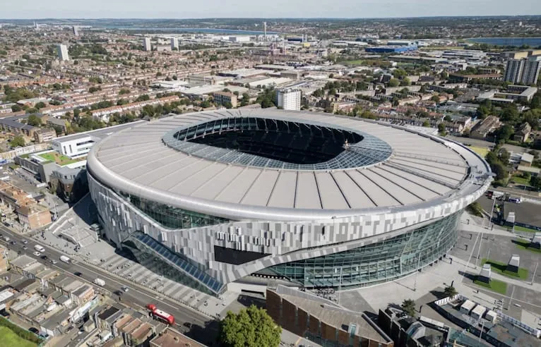 An aerial view of Tottenham Hotspur Stadium's curved glass and steel exterior, its retractable roof open, surrounded by the residential streets of N17