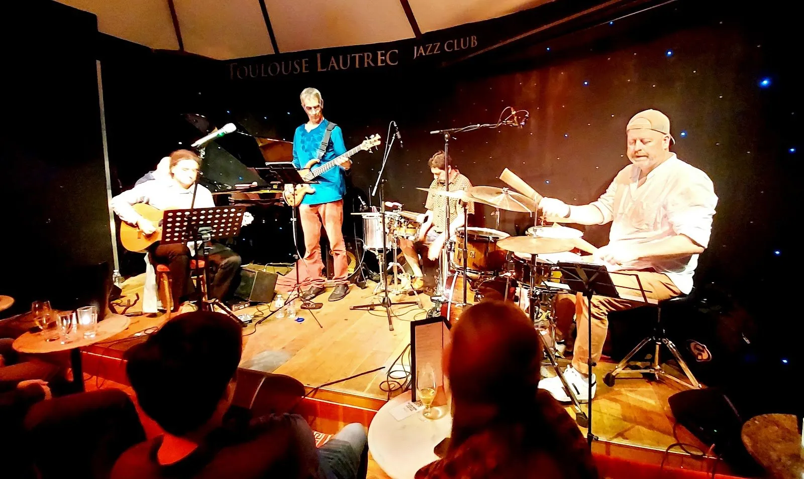 A four-piece band plays on the stage of the Toulouse Lautrec jazz club, with people seated at small tables in front