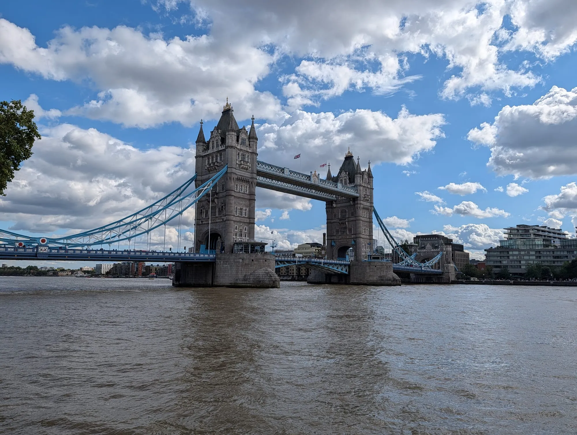 Tower Bridge seen along the Thames from the Tower's riverside wharf, both towers and the high walkway in full, under a bright sky
