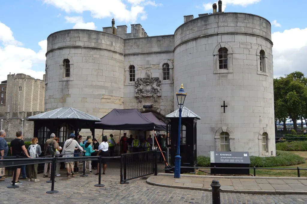Visitors queueing at the Tower of London's entrance beside the Middle Tower, with a bag-check marquee and a Yeoman Warder at the gate