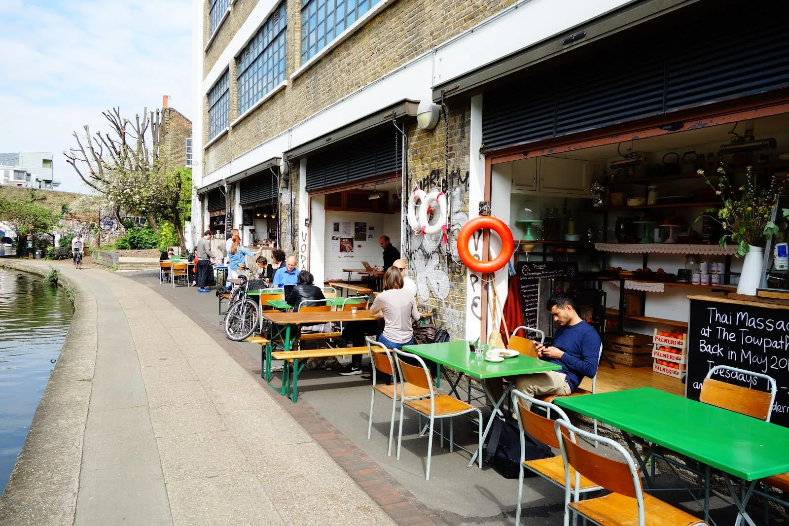 Towpath on the Regent's Canal: green folding tables and wooden chairs set out on the towpath in front of open shuttered units cut into the canal wall, with the counter, shelves of crockery and a chalkboard menu inside