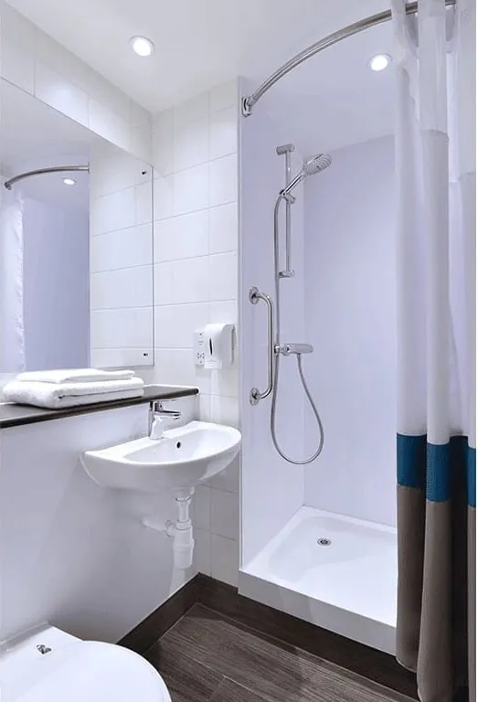 A small white bathroom at Travelodge London Central Marylebone with a shower tray behind a white and teal curtain, a hand shower on a rail and a small basin