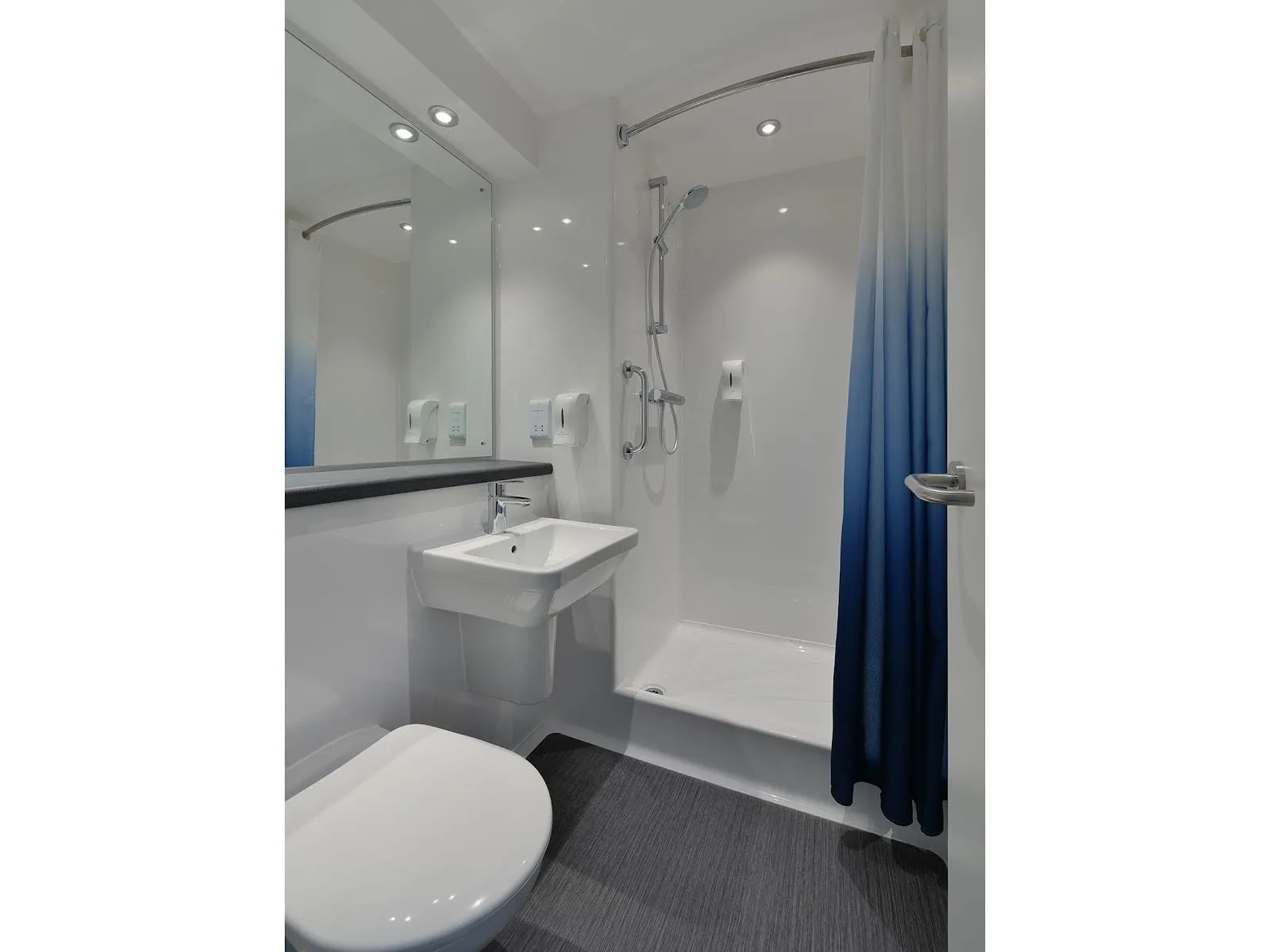 The ensuite at Travelodge London Docklands Central: white panelled walls, a shower tray behind a blue ombré curtain, a wall-hung basin and a wide mirror