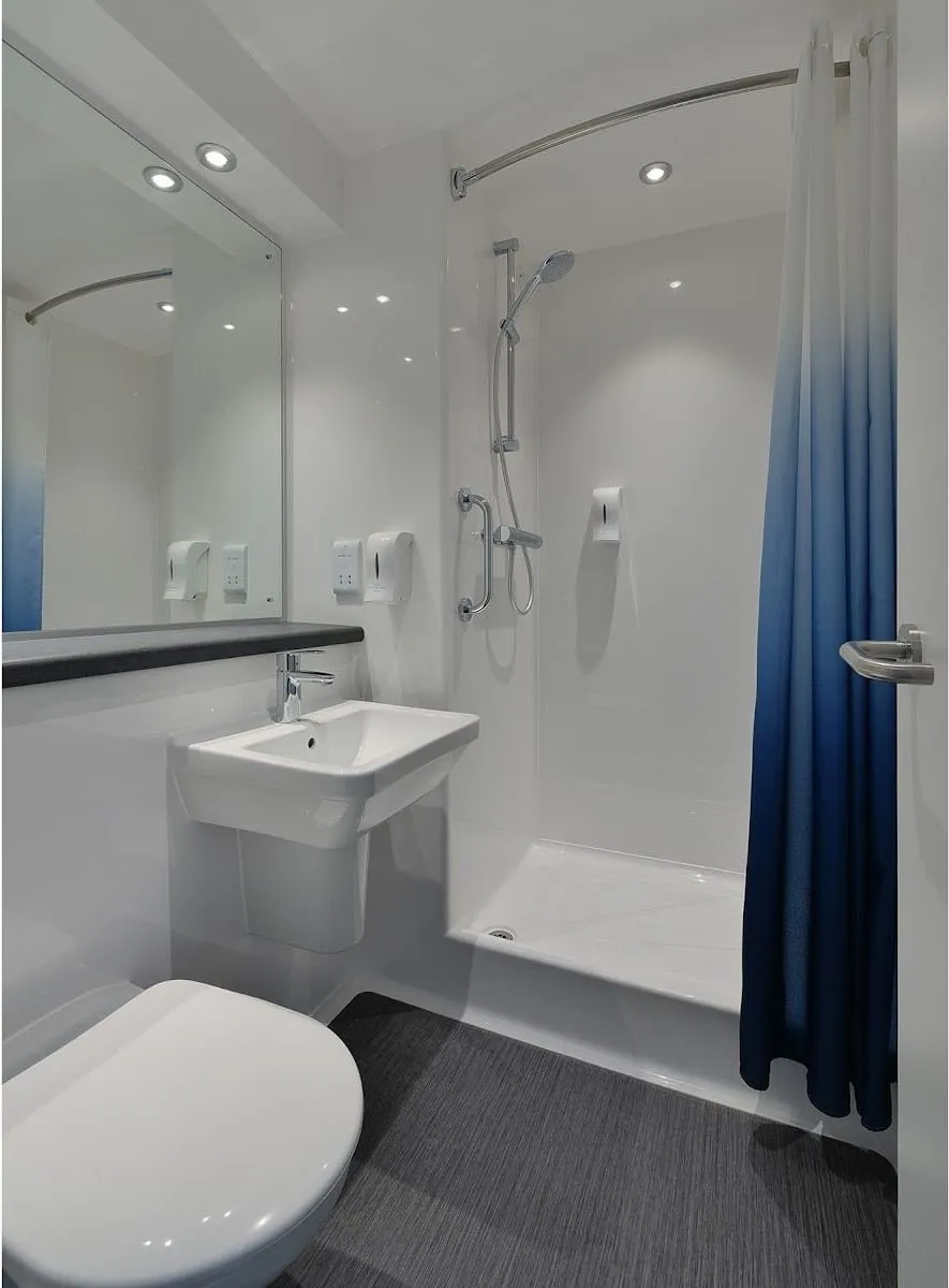 The shower room at Travelodge London Docklands Central, with a wall-hung basin and a shower behind a blue curtain