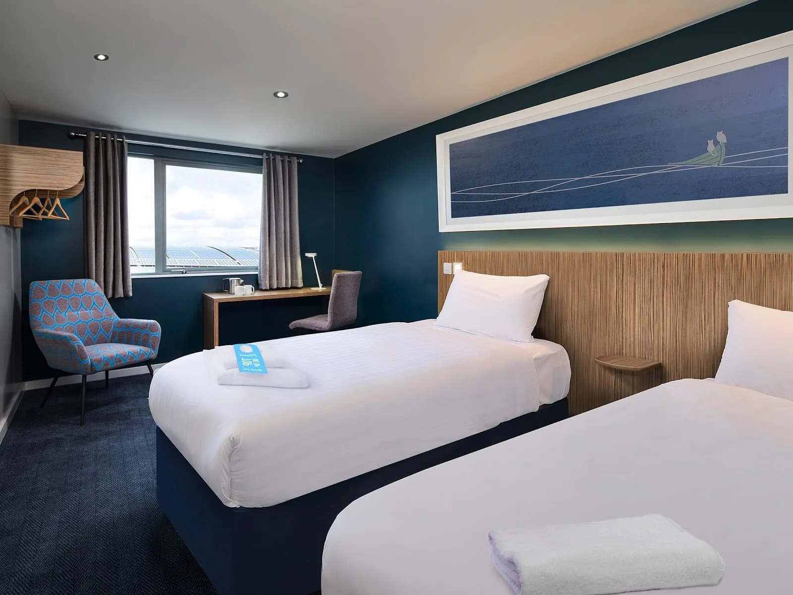 A twin room at Travelodge London Greenwich High Road with a blue wall, a patterned armchair and a desk by the window