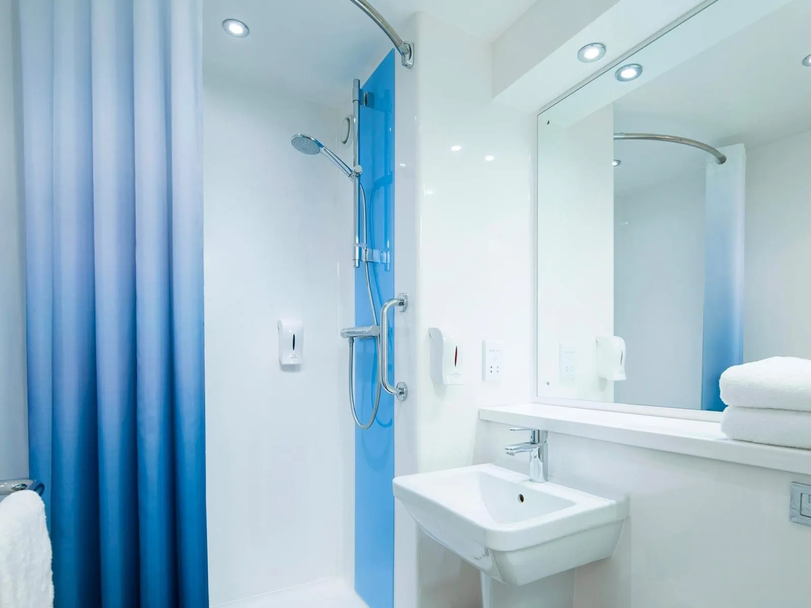 A white bathroom at Travelodge London Harrow with a walk-in shower, its hand shower on a rail, behind a blue ombré curtain and a square basin under a wide mirror