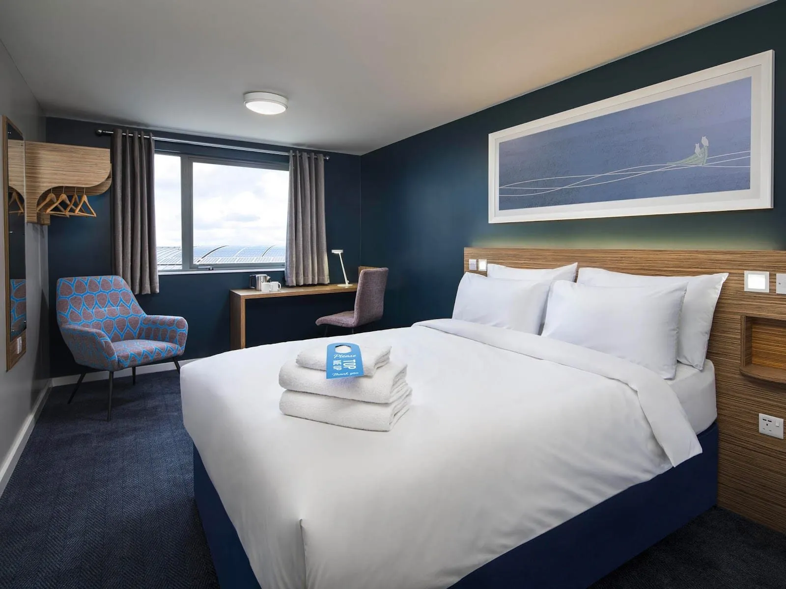 A double room at Travelodge London Harrow with a navy feature wall, a long framed print above a striped wood headboard, a patterned blue armchair and a desk under the window