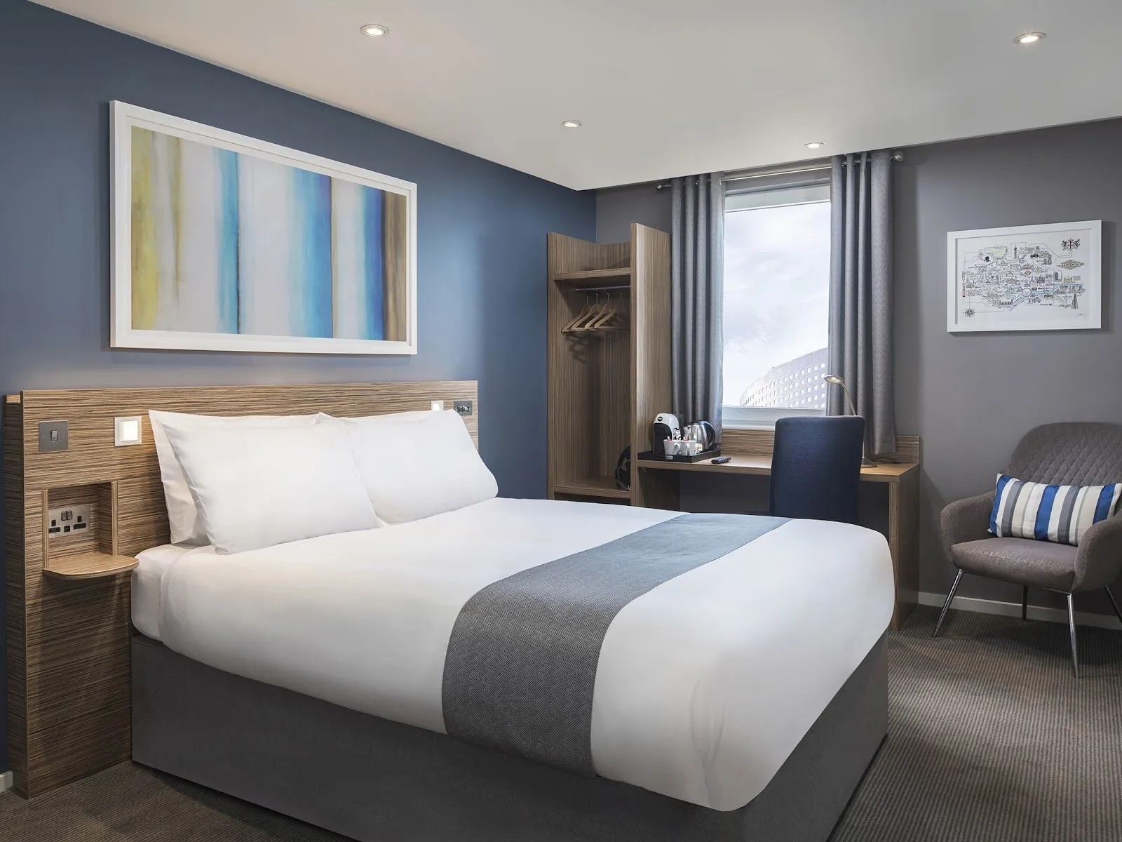 A typical Travelodge room, in navy and grey, with a wardrobe, desk and a framed London map on the wall