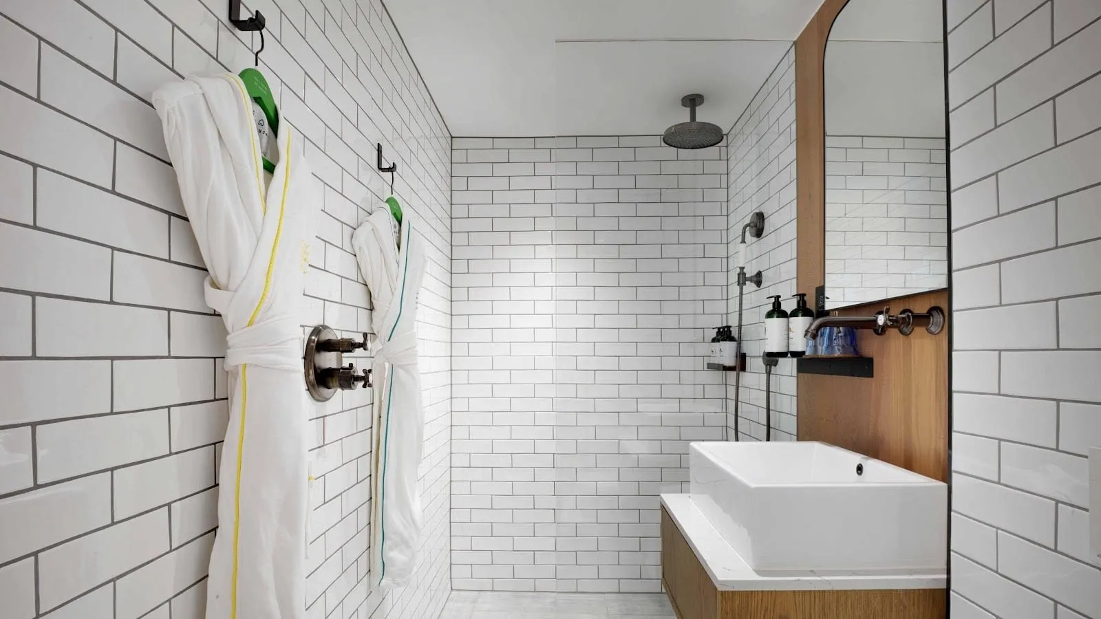 A Treehouse Hotel London bathroom in white metro tiles with grey grout, two bathrobes on hooks, a walk-in shower with a ceiling rain head and a square basin on an oak shelf